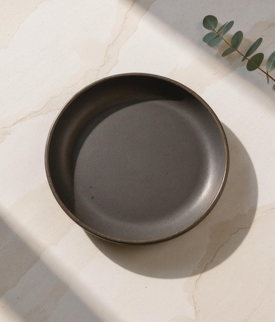 Danmero Charcoal Pasta Plate on pale travertine with sculptural shadow